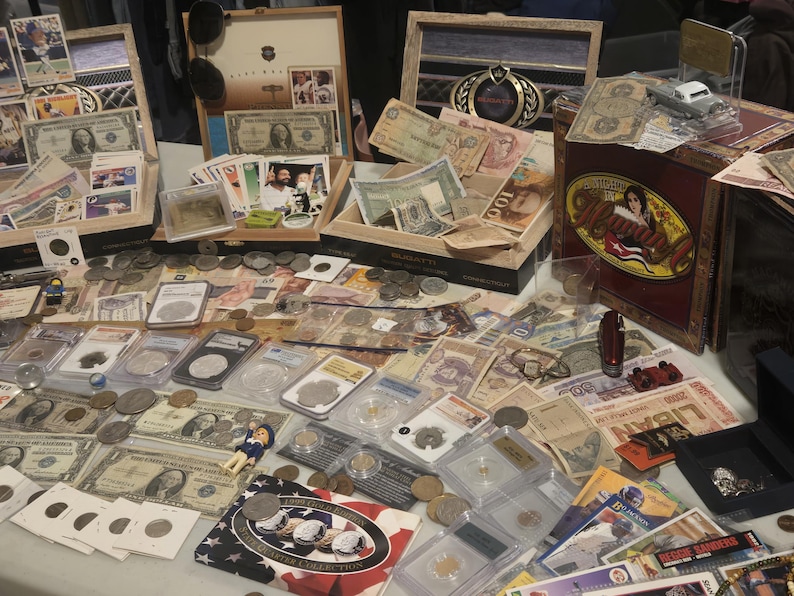 May include: A collection of vintage currency, coins, and collectibles displayed on a table. Items include paper money, coins in protective cases, trading cards, and a decorative cigar box. The items are arranged for sale.