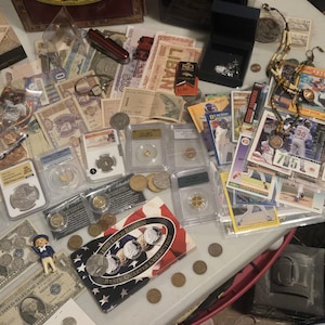 May include: A collection of vintage currency, coins, and collectibles, including paper money, baseball cards, and graded coins. The items are arranged on a table, showcasing a variety of historical and numismatic treasures.