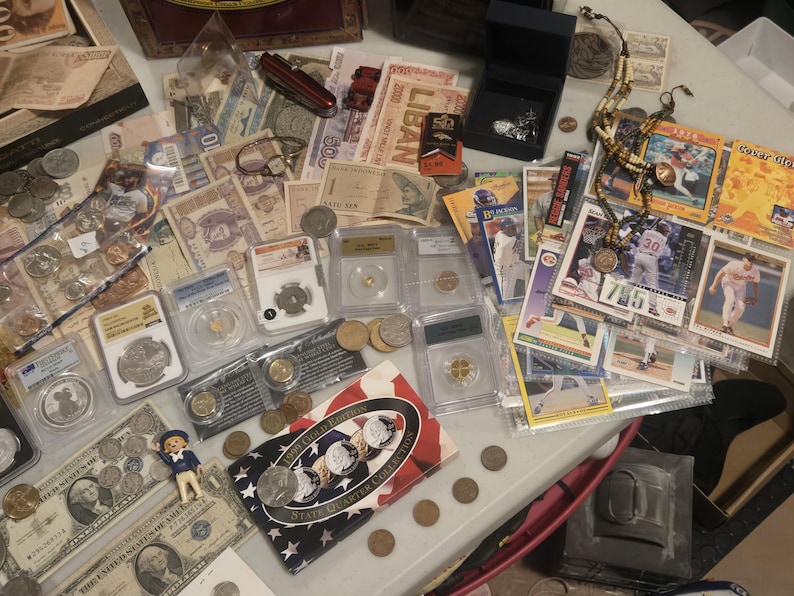 May include: A collection of vintage currency, coins, and collectibles, including paper money, baseball cards, and graded coins. The items are arranged on a table, showcasing a variety of historical and numismatic treasures.