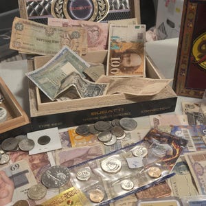 May include: A collection of vintage currency and coins, including paper bills and various coins. The items are displayed in wooden boxes and on a table, showcasing a variety of denominations and designs. The image includes the text "BUGATTI".