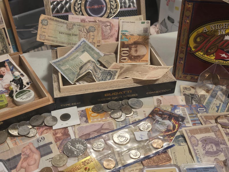 May include: A collection of vintage currency and coins, including paper bills and various coins. The items are displayed in wooden boxes and on a table, showcasing a variety of denominations and designs. The image includes the text "BUGATTI".