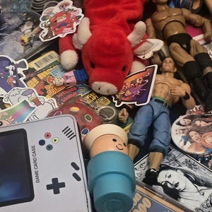 May include: A collection of assorted toys and memorabilia including a red plush bull, several action figures, a light gray game card case, a blue plastic figure, a Backstreet Boys Golden Hits 2000 CD, various stickers, and a small pink plastic suitcase.