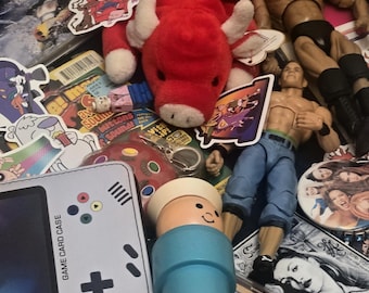 Puede incluir: Una colección de juguetes y recuerdos variados que incluye un toro de peluche rojo, varias figuras de acción, un estuche para tarjetas de juego gris claro, una figura de plástico azul, un CD de Backstreet Boys Golden Hits 2000, varias pegatinas y una pequeña maleta rosa.