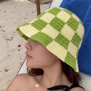 Puede incluir: Un sombrero de pescador tejido a ganchillo con un patrón de tablero de ajedrez en verde claro y crema. El sombrero lo lleva una persona, con el ala proyectando una sombra. Un accesorio de verano.