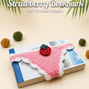 Può includere: Un segnalibro all'uncinetto con una base triangolare rosa, un bordo smerlato bianco e una piccola decorazione a forma di fragola rossa con foglie verdi. Il testo Strawberry Bookmark PDF Crochet Pattern è visualizzato sopra.