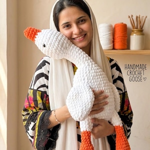 Può includere: Un'oca all'uncinetto fatta a mano in bianco con becco e zampe arancioni. Il peluche ha occhi di sicurezza neri. Il testo "HANDMADE CROCHET GOOSE" è visibile nell'immagine.