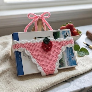 Könnte beinhalten: Ein rosa Häkel-Lesezeichen in Form von Unterwäsche mit weißen, gewellten Kanten und einer roten Erdbeere. Das Lesezeichen liegt auf einem Buch, im Hintergrund sind Erdbeeren und ein Notizbuch zu sehen.