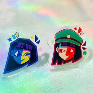 May include: Two clear acrylic pins featuring stylized character portraits. One pin has a blue and yellow face with black hair, the other has a red and black face with a green hat. Both pins have star and geometric accents.