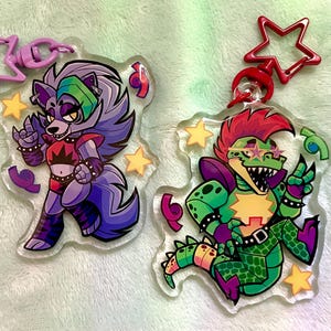 Puede incluir: Dos llaveros de acrílico transparente con personajes de dibujos animados. Uno es un lobo morado y gris con una parte superior roja, el otro es un caimán verde con una cresta roja. Ambos tienen cierres en forma de estrella y estrellas amarillas.