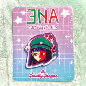 Peut inclure: Une &eacute;pingle en acrylique de 3,8 cm repr&eacute;sentant un personnage stylis&eacute; avec un chapeau vert, des cheveux rouges et noirs et un visage rouge et blanc. L'&eacute;pingle est sur une carte avec le texte "ENA" en vert et rouge, et "1.5" Acrylic Pin". La carte a un motif de grille et le texte "By Scruffy Shoppe".