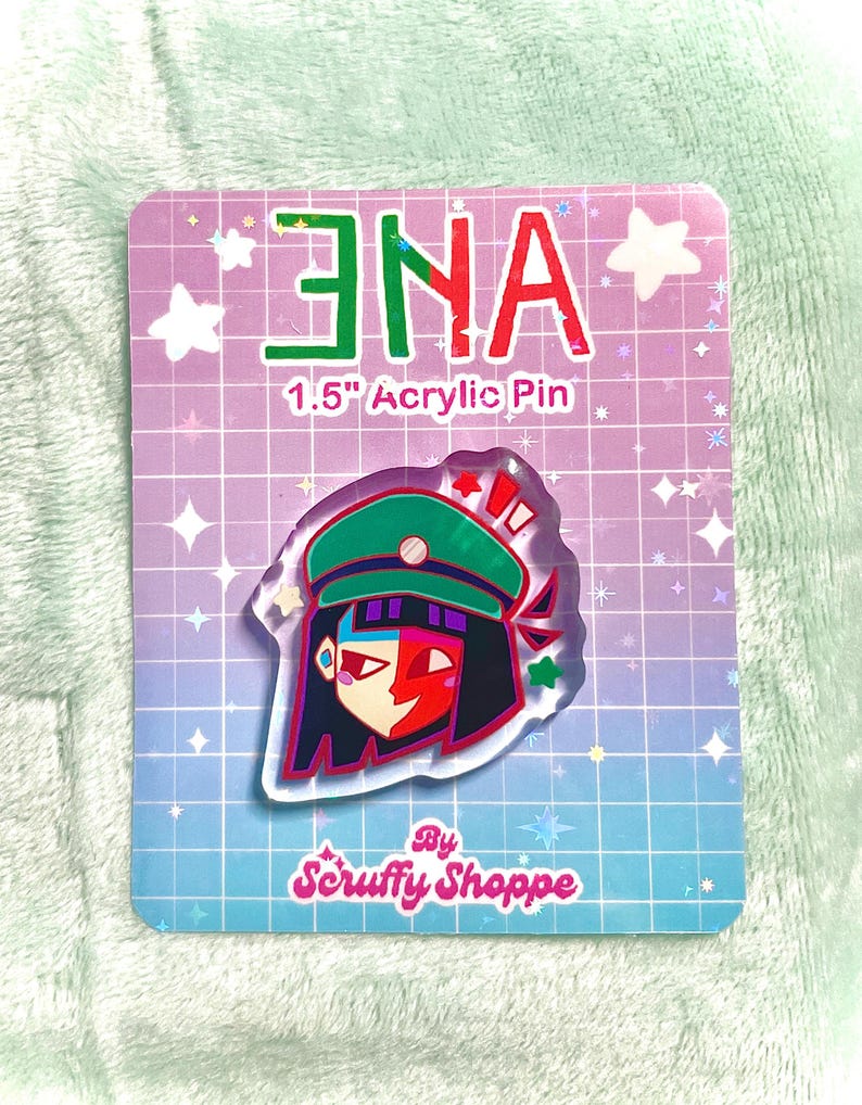 Peut inclure: Une &eacute;pingle en acrylique de 3,8 cm repr&eacute;sentant un personnage stylis&eacute; avec un chapeau vert, des cheveux rouges et noirs et un visage rouge et blanc. L'&eacute;pingle est sur une carte avec le texte "ENA" en vert et rouge, et "1.5" Acrylic Pin". La carte a un motif de grille et le texte "By Scruffy Shoppe".