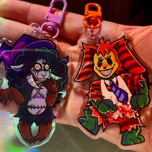 Op de afbeelding: Twee acryl sleutelhangers met cartoonfiguren. Eén is een donkerharig wezen met hoorns en een gestikte torso. De andere is een rood-oranje clownachtig figuur met een brede grijns en een gestreepte stropdas. Beide hebben kleurrijke accenten.