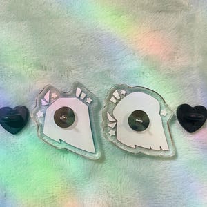 Peut inclure: Deux &eacute;pingles en acrylique transparent avec un motif blanc et des &eacute;toiles, ainsi que deux fermoirs noirs en forme de c&oelig;ur. Les &eacute;pingles ont un dos argent&eacute; et sont pos&eacute;es sur un fond aux couleurs de l'arc-en-ciel.