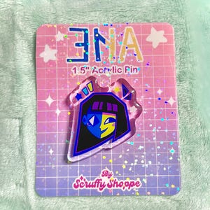 Peut inclure: &Eacute;pingle en acrylique transparente repr&eacute;sentant un personnage de dessin anim&eacute; avec une coupe au carr&eacute; noire, un visage bleu et jaune, et un contour noir. L'&eacute;pingle est sur une carte rose et violette avec le texte "ENA" et "1.5" Acrylic Pin". La carte porte &eacute;galement le texte "By Scruffy Shoppe".