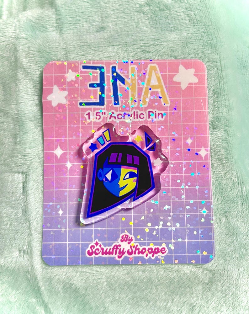 Peut inclure: &Eacute;pingle en acrylique transparente repr&eacute;sentant un personnage de dessin anim&eacute; avec une coupe au carr&eacute; noire, un visage bleu et jaune, et un contour noir. L'&eacute;pingle est sur une carte rose et violette avec le texte "ENA" et "1.5" Acrylic Pin". La carte porte &eacute;galement le texte "By Scruffy Shoppe".