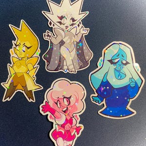 May include: Four holographic stickers featuring cartoon characters. The characters include a yellow figure, a white figure with a cape, a pink figure, and a blue figure. Each sticker has a unique design and pose, with a sparkly, iridescent finish.
