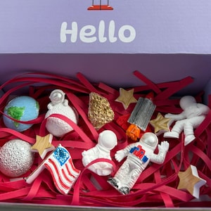 Puede incluir: Una caja lila con la palabra "Hello" en blanco, que contiene figuras con temática espacial y tiras de papel rojo. Las figuras incluyen astronautas, un globo terráqueo, una luna, estrellas, una pepita de oro y una bandera estadounidense.