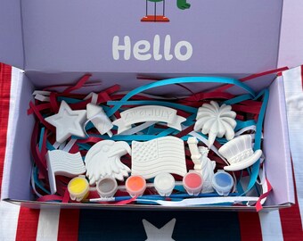 May include: Open box with a patriotic theme. Inside, there are star, flag, and eagle-shaped items, along with small paint pots. The box is lined with red, white, and blue paper strips. The box lid has a cartoon bird and the word "Hello".