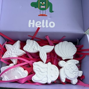 Puede incluir: Una caja lila con un personaje de dibujos animados y la palabra "Hello". Contiene figuras blancas de criaturas marinas: estrella de mar, pez, concha, caballito de mar y tortuga, dispuestas en papel triturado rosa.