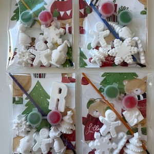 Puede incluir: Cuatro bolsas de plástico transparente que contienen kits de manualidades navideñas para pintar. Cada kit incluye figuras de cerámica blanca como hombres de jengibre, copos de nieve y árboles de Navidad, junto con botes de pintura y un pincel. El fondo presenta un diseño festivo de oso y árbol.
