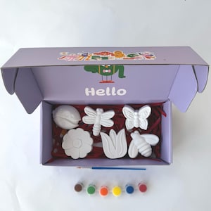 Puede incluir: Una caja morada con la palabra "Hello" en la parte delantera, abierta para revelar un conjunto de seis figuritas de cerámica blanca de una mariquita, una libélula, una mariposa, una flor, un tulipán y una abeja. La caja también contiene un pincel y seis pequeños botes de pintura en marrón, verde, naranja, rojo, amarillo y azul.