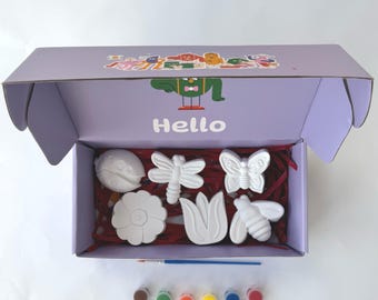 May include: A purple box with the word "Hello" on the front, open to reveal a set of six white ceramic figurines of a ladybug, dragonfly, butterfly, flower, tulip, and bee. The box also contains a paintbrush and six small paint pots in brown, green, orange, red, yellow, and blue.