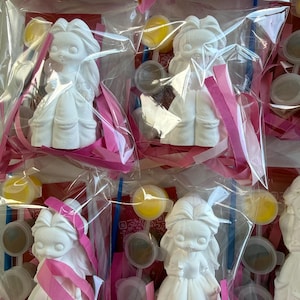 May include: Several paint-your-own ceramic figurines of a princess in a clear plastic bag with pink ribbon and paint supplies. The figurines are white and have long hair and a dress. Yellow paint and paintbrushes are also visible.