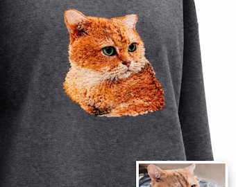 Puede incluir: Sudadera gris oscuro con un bordado de un gato naranja con ojos verdes. El gato es el punto focal, con pelaje detallado y una expresión pensativa. Una imagen más pequeña del gato está en la esquina inferior derecha.
