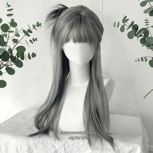 Puede incluir: Una peluca larga y gris con un estilo en capas, con flequillo y cola de caballo. La peluca se muestra en una cabeza de maniquí. El texto "Justsomestuff4you wig for you" es visible en la parte inferior.