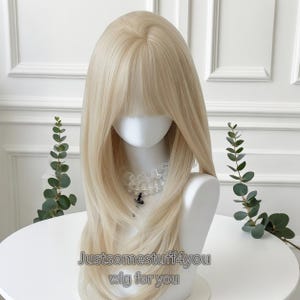 May include: A light blonde wig with long, layered hair and bangs, displayed on a mannequin head. The wig has a natural-looking hairline and a soft, flowing texture. The text "Justsomestuff4you wig for you" is visible at the bottom of the image.