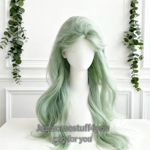 Puede incluir: Una peluca larga y ondulada en un suave color verde apagado. La peluca está peinada con ondas sueltas y una raya de aspecto natural. El texto "Justsomestuff4you wig for you" es visible en la parte inferior. La peluca se muestra en una cabeza de maniquí blanca.