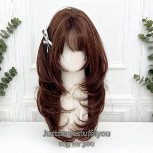 Puede incluir: Peluca marrón a capas con flequillo, peinada sobre una cabeza de maniquí. Un pequeño lazo blanco está prendido a un lado. El texto "Justsomestuff4you wig for you" está impreso en la parte inferior.