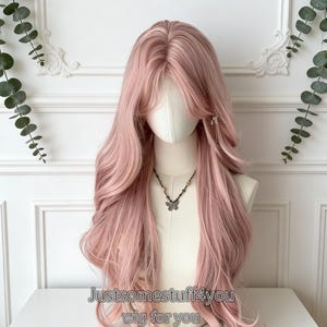 Può includere: Una parrucca lunga e ondulata rosa chiaro, acconciata su una testa di manichino. La parrucca ha una scriminatura naturale e strati, con il testo "Justsomestuff4you wig for you" in basso. È visibile una collana con pendente a farfalla.