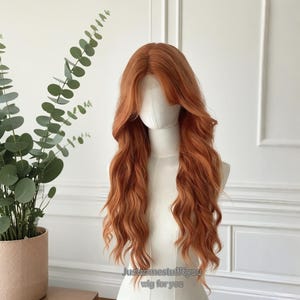 Może przedstawiać: Długa, falista peruka w kolorze kasztanowym na głowie manekina. Peruka ma przedziałek z boku i grzywkę, z opadającymi lokami. Manekin ustawiony jest na białym tle, z rośliną w doniczce po lewej stronie.