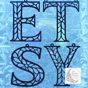 May include: Black laser-cut wooden letters spelling "ETSY" with decorative leaf-like patterns inside each letter. Each letter has a small hole at the top for hanging. The background is a light blue, textured surface. A small logo is in the bottom right corner.