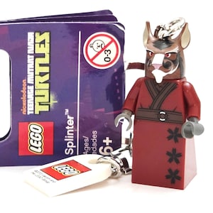 May include: A LEGO Teenage Mutant Ninja Turtles Splinter keychain featuring a brown rat-like character with a white beard, wearing a dark red robe with black floral patterns. The attached purple packaging displays the text LEGO Teenage Mutant Ninja Turtles.