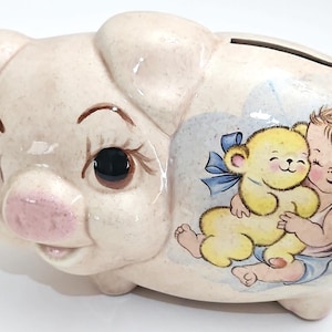 Può includere: Un salvadanaio in ceramica a forma di maiale, dipinto in una tonalità rosa chiaro. La banca presenta un'illustrazione dipinta di un bambino che abbraccia un orsacchiotto giallo. Il salvadanaio ha una fessura per le monete sulla parte superiore.