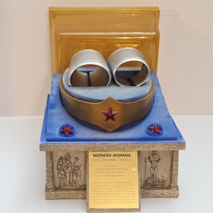 May include: A gold-colored tiara with a red star, two silver-colored bracelets, and a display base. The base features blue velvet, two small red stars, and a gold plaque with the text WONDER WOMAN Tiara / Bracelets / Earrings and museum details.