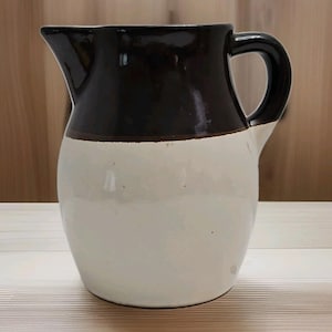 May include: A ceramic pitcher featuring a two-tone design with a glossy dark brown glaze on the upper portion and a smooth off-white glaze on the lower body. The vessel includes a curved handle on the right side and a distinct pouring spout.