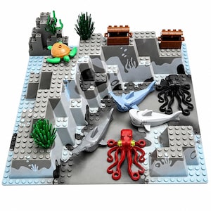 May include: A building block playset featuring an underwater scene with gray rock formations, two brown treasure chests, a green turtle, a black octopus, a red octopus, a light blue shark, a white shark, a gray sawfish, and green seaweed plants.