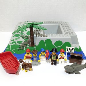 May include: A Lego pirate-themed playset featuring a red rowboat, a gray island base with a palm tree, and several pirate minifigures. Includes a treasure chest, a shark, and a monkey figurine. The set has a pirate flag.
