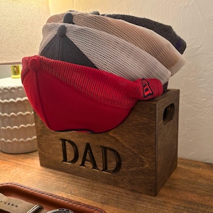 May include: A wooden hat rack with the word "DAD" engraved on the front. The rack is filled with four baseball caps, one red, one white, one beige, and one dark blue.