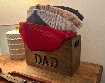 Puede incluir: Un perchero de madera para sombreros con la palabra "DAD" grabada en la parte delantera. El perchero está lleno de cuatro gorras de béisbol, una roja, una blanca, una beige y una azul oscuro.