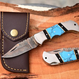 May include: A set of pocket knives with turquoise handles and a brown leather sheath. The knives have silver blades with intricate patterns. A sharpening tool is included.