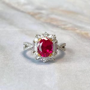 Może przedstawiać: Srebrny pierścionek z dużym, owalnym rubinem w centrum. Rubinu otacza aureola z mniejszych, przezroczystych kamieni szlachetnych i złotych akcentów. Na obrączce pierścionka znajdują się dodatkowe, przezroczyste kamienie.