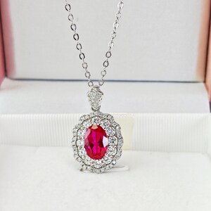 May include: A silver necklace with a large oval ruby pendant. The ruby is surrounded by a halo of small, sparkling white gemstones. The pendant hangs from a delicate silver chain, displayed in a white jewelry box.