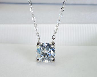 Puede incluir: Un collar de plata con un gran colgante de piedra preciosa transparente y de corte redondo. La piedra preciosa está sujeta por cuatro garras y cuelga de una delicada cadena. El colgante brilla, reflejando la luz. El fondo es blanco suave.