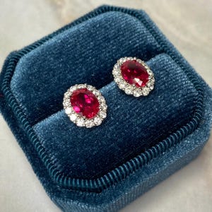 May include: A pair of oval ruby and diamond earrings in a blue velvet jewelry box. The earrings feature a red ruby surrounded by a halo of small diamonds.