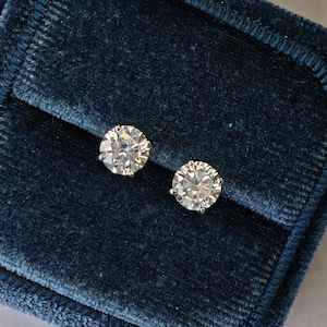 Può includere: Due orecchini a lobo con diamanti taglio rotondo. I diamanti sono incastonati in un metallo color argento e sono presentati su una superficie di velluto blu scuro. Gli orecchini brillano e riflettono la luce.