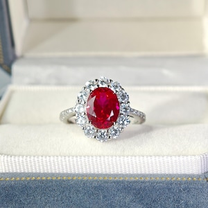 May include: A silver-toned ring featuring a large, oval-cut vibrant red gemstone at the center, surrounded by a halo of small, clear, round-cut white stones. The band is also set with small, clear, round-cut white stones along the visible sides.